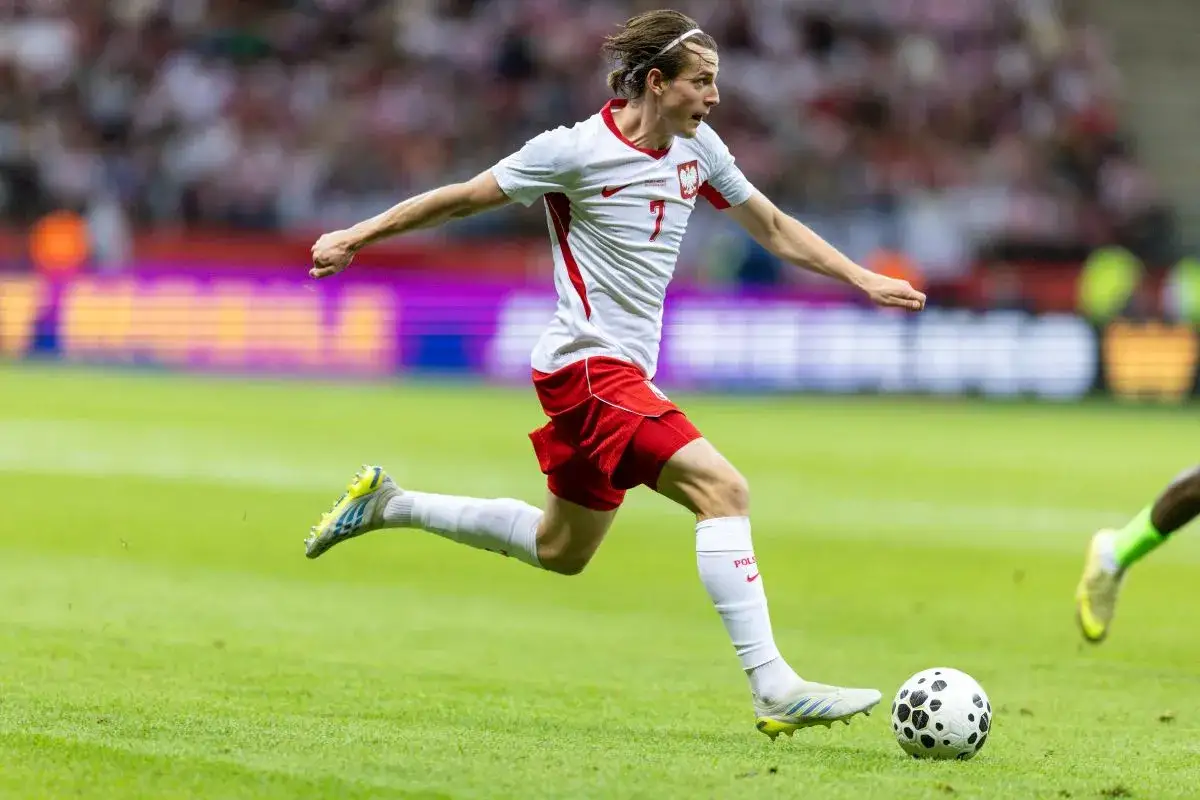 Jakub Kamiński w akcji, biegnie z piłką na boisku. Jego obecne drużyny to reprezentacja Polski i VfL Wolfsburg.