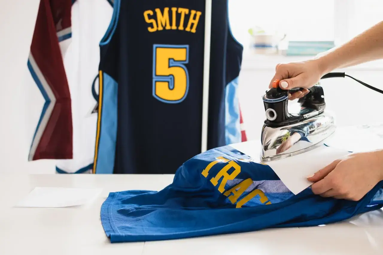 Une main utilise un fer &agrave; repasser pour d&eacute;froisser un maillot de basket-ball bleu avec des lettres jaunes. Un autre maillot bleu marine avec le nom 