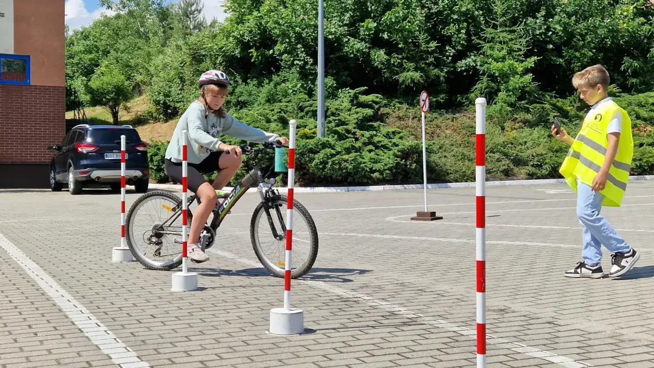 Dziewczynka na rowerze ćwiczy jazdę między pachołkami. Czy karta rowerowa jest obowiązkowa? Chłopiec w kamizelce obserwuje.