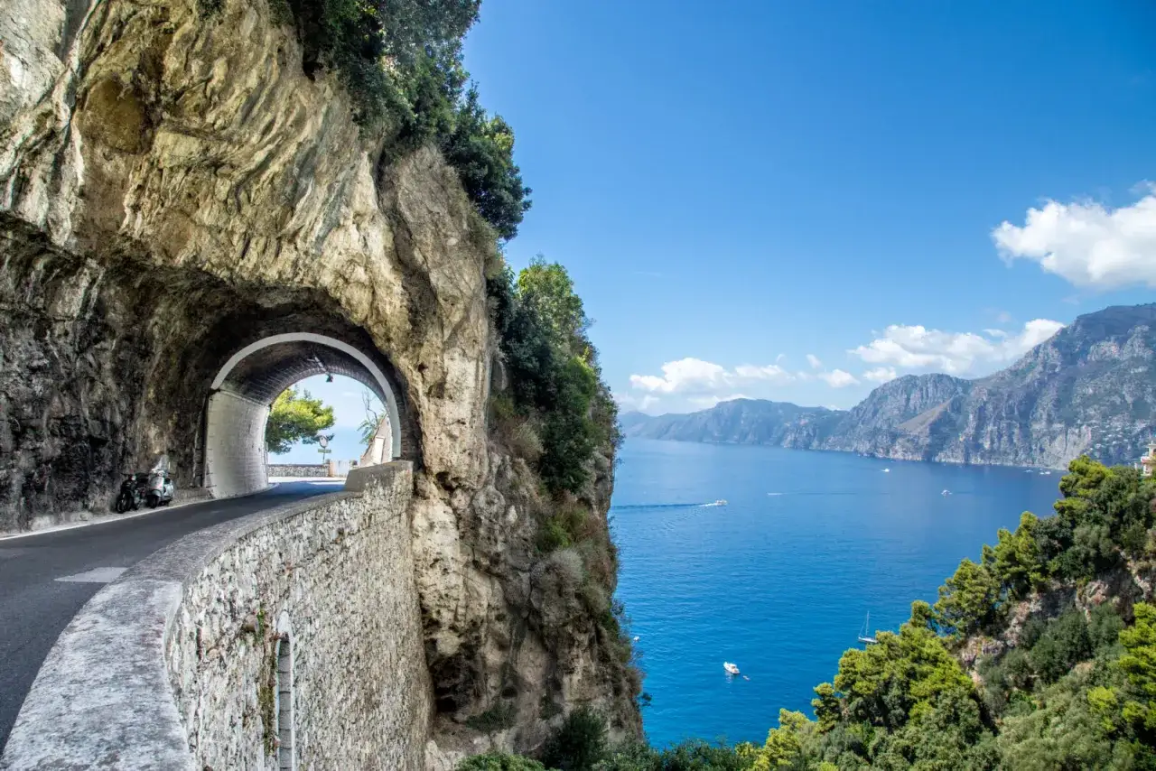 A scenic tunnel on a coastal road, perfect for an Italy road trip, opens to a stunning view of the blue sea and mountains.