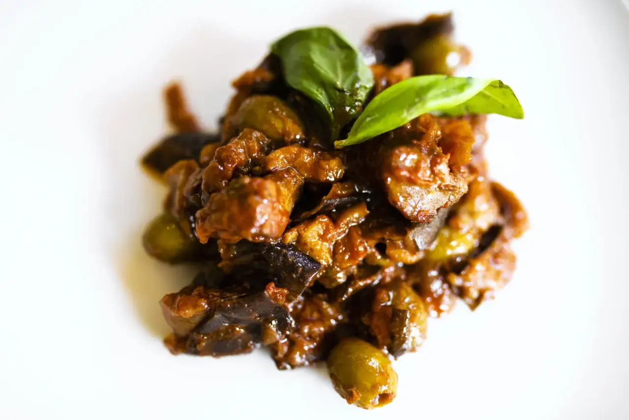 Un saporito piatto di caponata di pesce spada, con melanzane, olive e basilico fresco, su uno sfondo bianco.