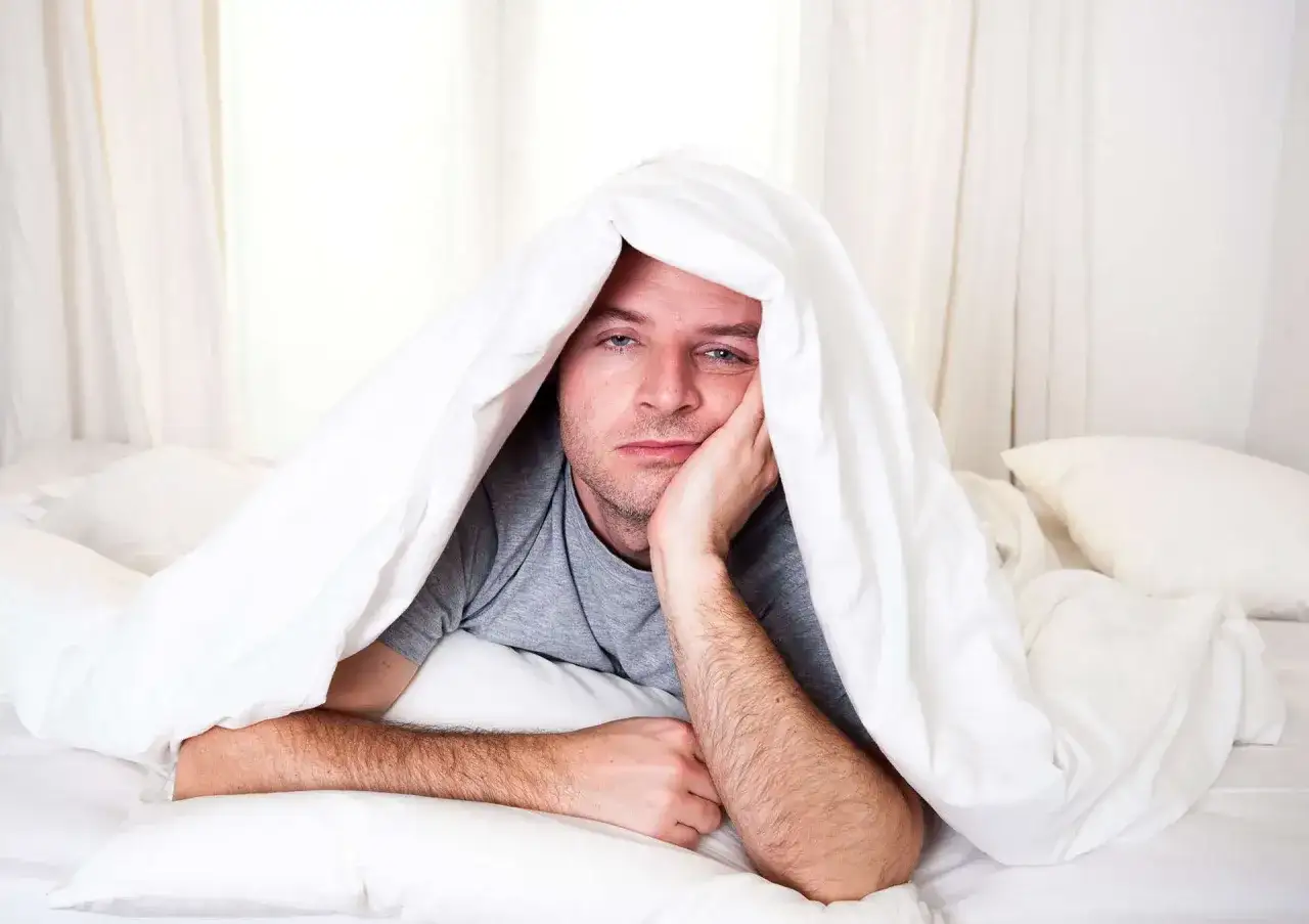 Homme au lit, sous une couette blanche, l'air fatigué, il a vraiment du mal à avoir les yeux en face des trous ce matin.