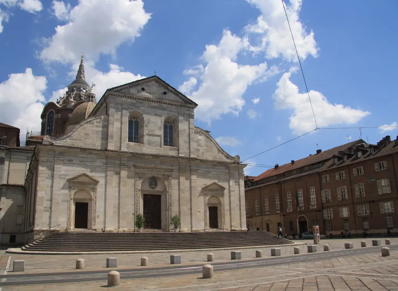 Il Duomo di Torino, con la sua imponente facciata in pietra e la cupola maestosa, si staglia sotto un cielo azzurro punteggiato di nuvole bianche.