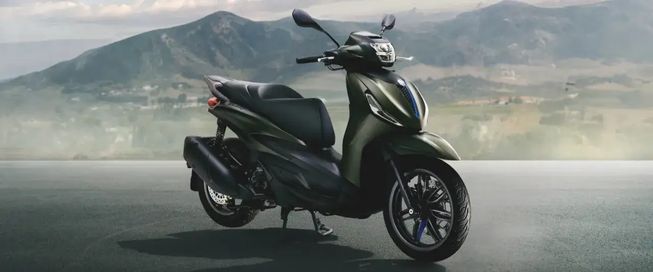 Oliwkowy Piaggio Beverly z niebieskimi akcentami na tle górskiego krajobrazu.