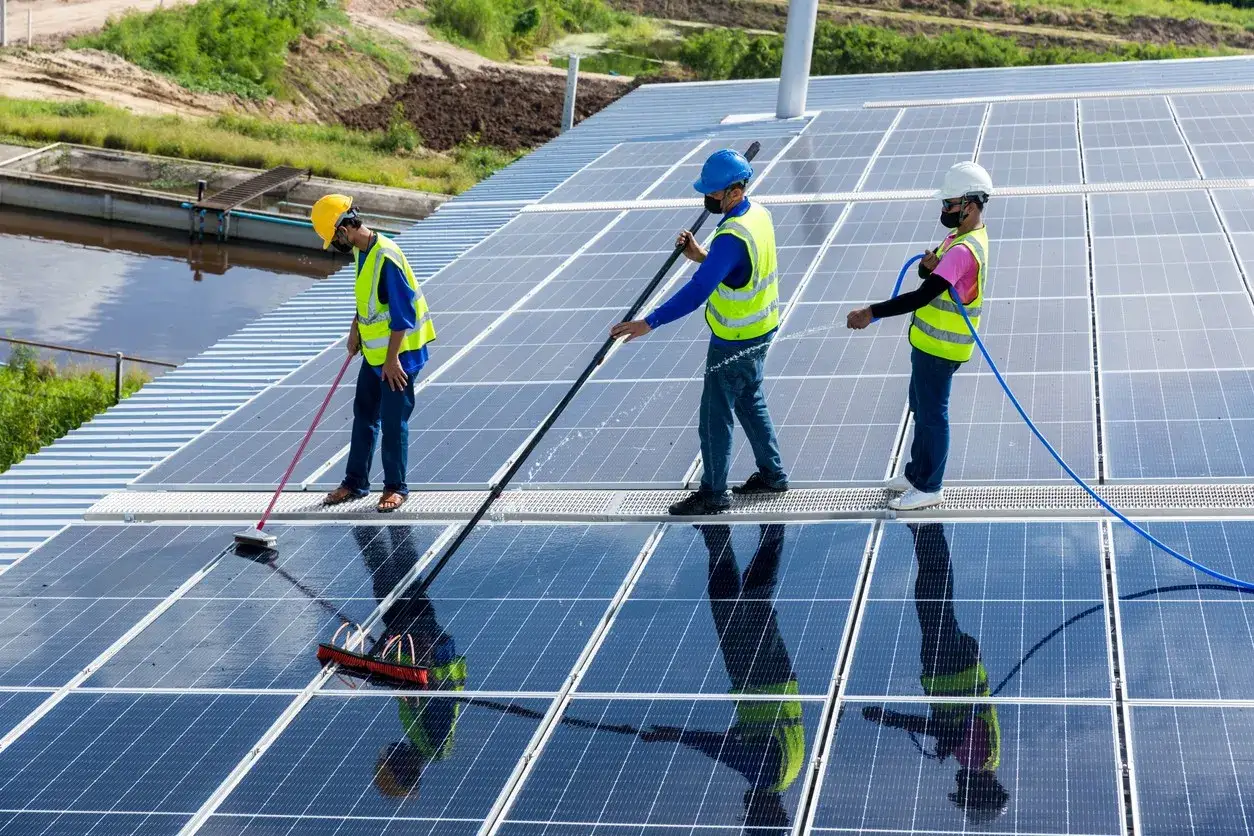 Drei Arbeiter reinigen eine Solaranlage auf einem Dach. Sie verwenden B&uuml;rsten und Wasser, um die Panels f&uuml;r optimale Leistung zu s&auml;ubern.