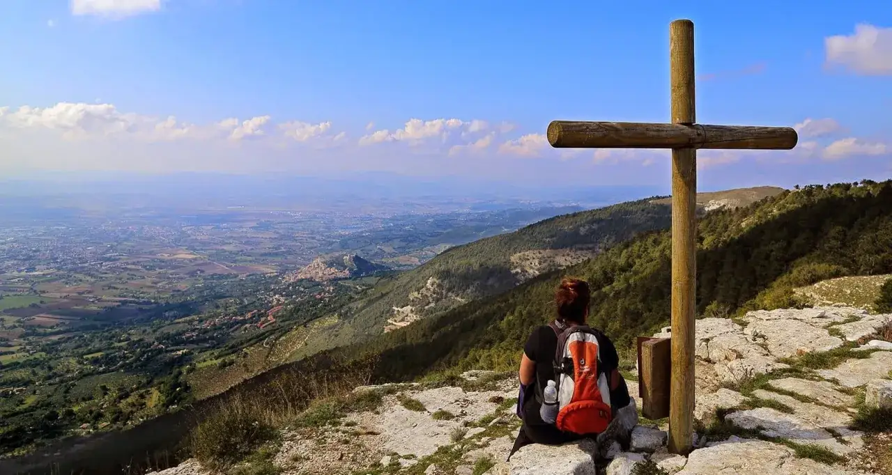 Vista panoramica dal monte Subasio, con una croce in primo piano e una persona che ammira la valle sottostante.