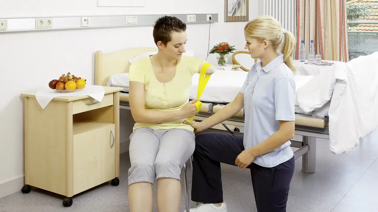 Rehabilitacja po amputacji ręki. Pacjentka ćwiczy z fizjoterapeutką, używając ż&oacute;łtej gumy oporowej.