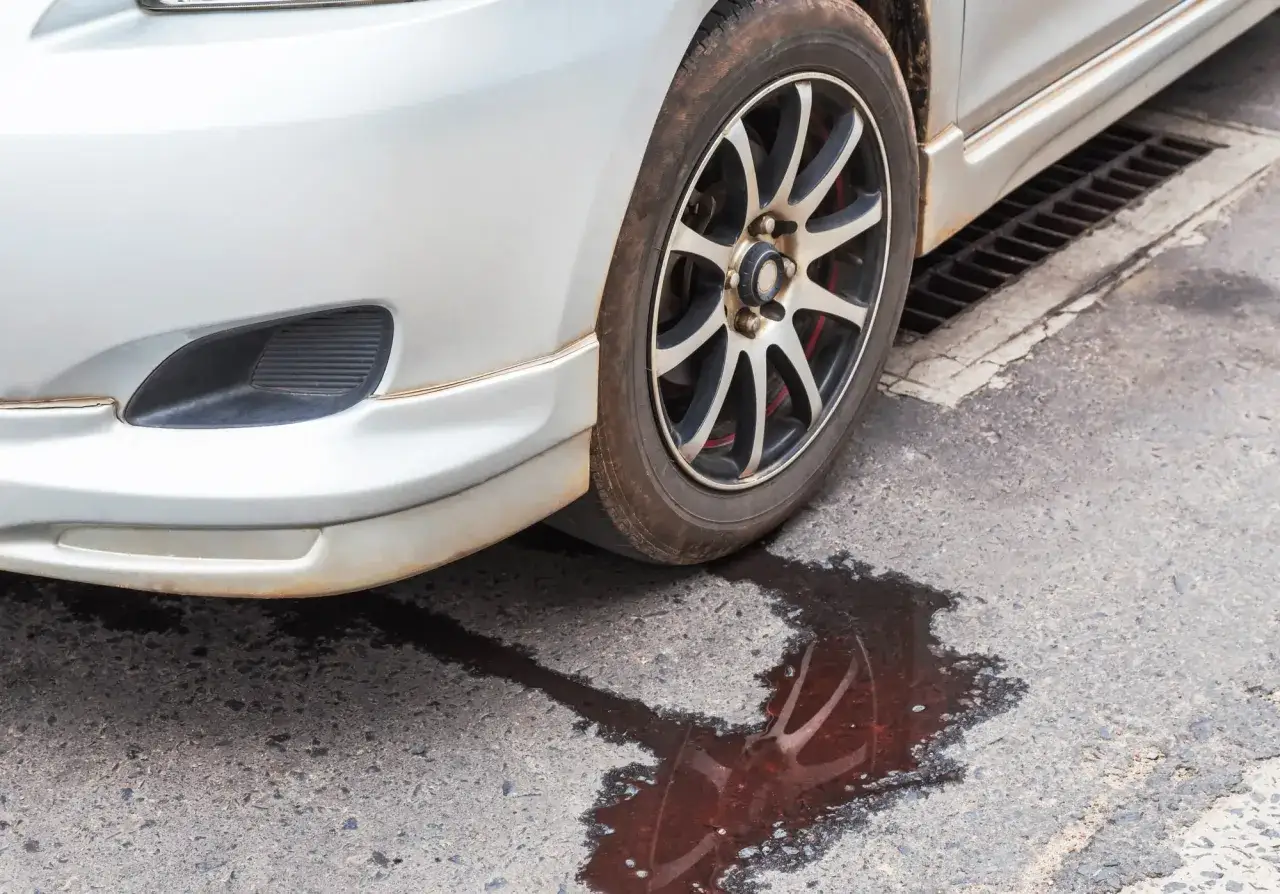 En silverf&auml;rgad bil st&aring;r parkerad vid en gatubrunn. En r&ouml;daktig v&auml;tska syns som droppar under bilen, vilket tyder p&aring; ett l&auml;ckage.