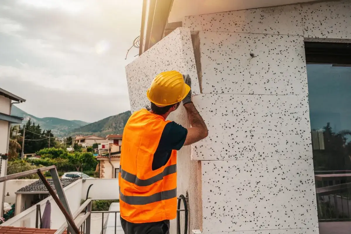 Operaio con casco giallo e giubbotto arancione applica pannelli isolanti per il cappotto casa.
