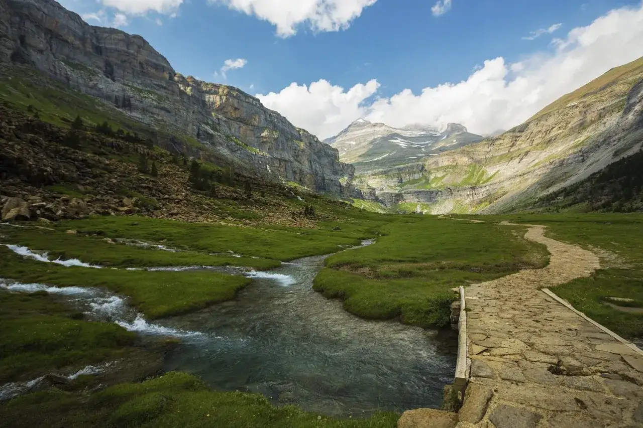 Un arroyo serpentea por un prado verde hacia imponentes montañas. Un camino de piedra cruza el agua, invitando a explorar estos impresionantes espacios naturales España.