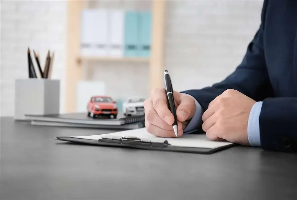Un uomo in giacca e cravatta firma un documento. Sul tavolo, modelli di auto e matite. L'assicurazione auto deve essere intestata al proprietario.