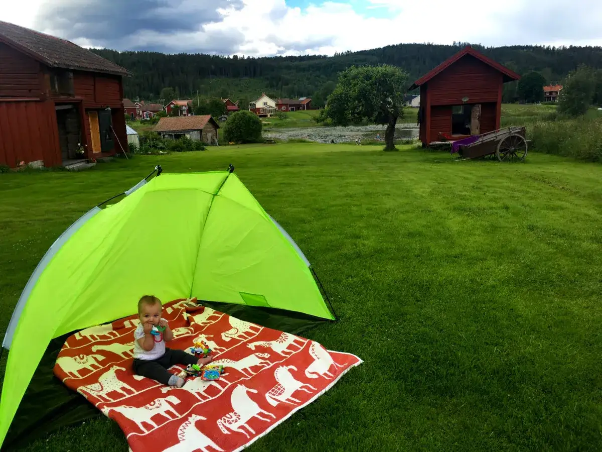 Liten bebis njuter av sol och lek p&aring; en r&ouml;d filt under ett gr&ouml;nt solskydd. I bakgrunden syns r&ouml;da hus och en vacker svensk landsbygd.