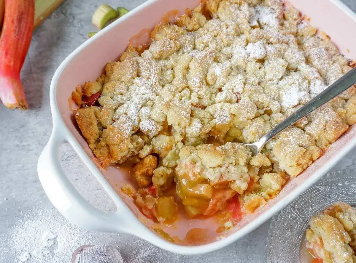 Ein frisch gebackener Rhabarber Crumble in einer Auflaufform, best&auml;ubt mit Puderzucker. Ein L&ouml;ffel steckt in der s&uuml;&szlig;en Leckerei.