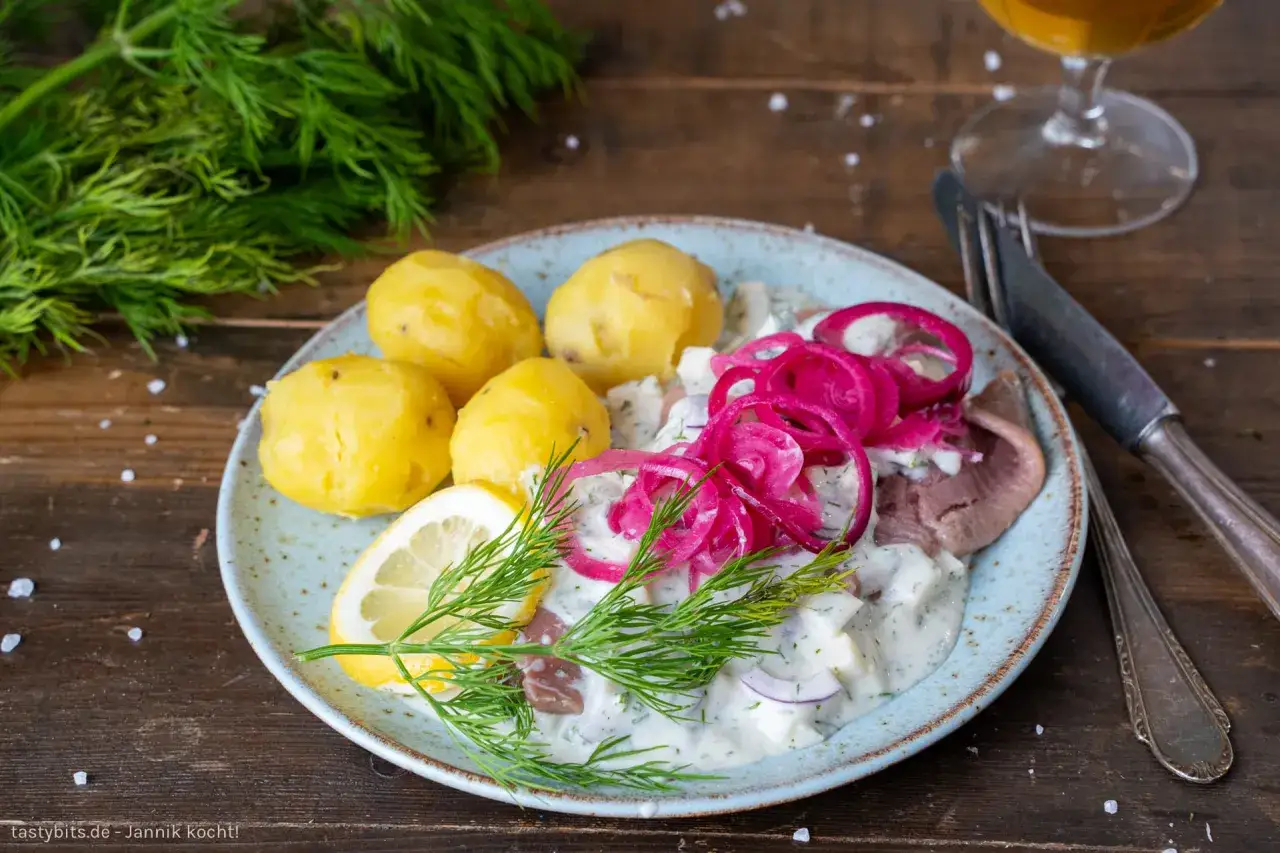 Matjes mit Bratkartoffeln: Ein Teller mit pochierten Kartoffeln, Matjesfilet in cremiger Sauce, roten Zwiebelringen, Zitrone und Dill.