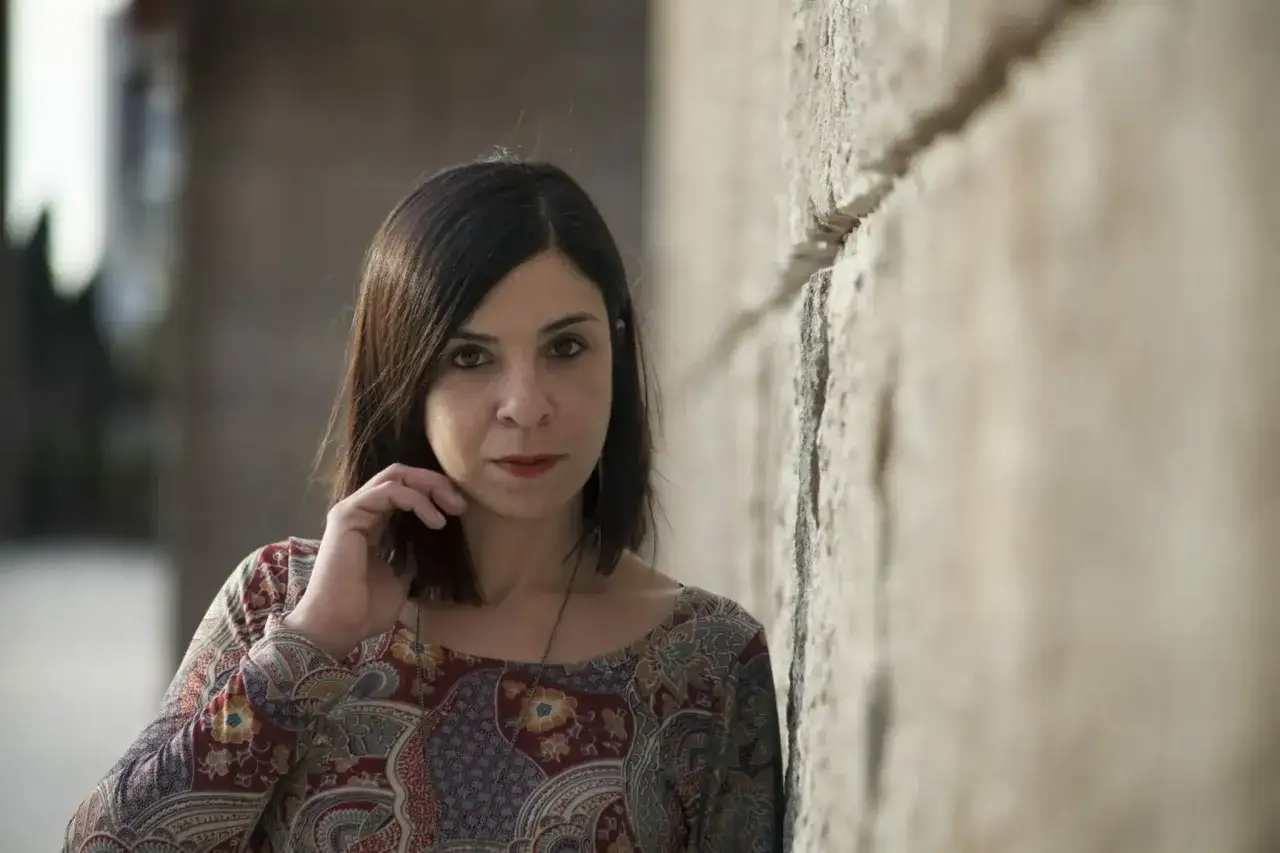 Una donna con capelli scuri e un vestito colorato appoggiata a un muro di pietra. Il suo sguardo intenso sembra raccontare una storia nera.