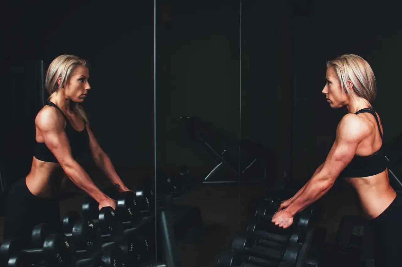 Eine **bodybuilder frau** greift nach Hanteln im Fitnessstudio. Ihr starker Körper spiegelt sich im dunklen Spiegel.