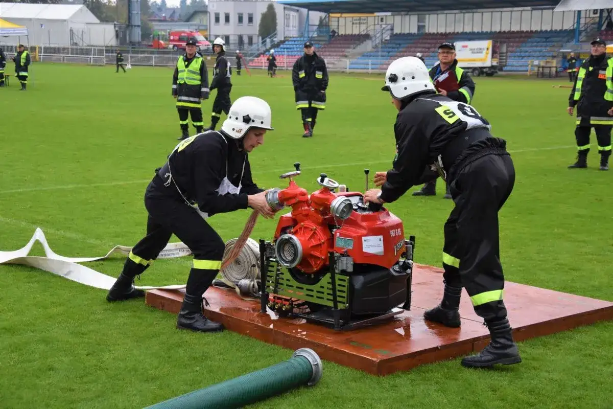 Strażacy przygotowują sprzęt do zawodów OSP. Dwie osoby w hełmach i czarnych strojach z żółtymi pasami pracują przy czerwonej pompie.