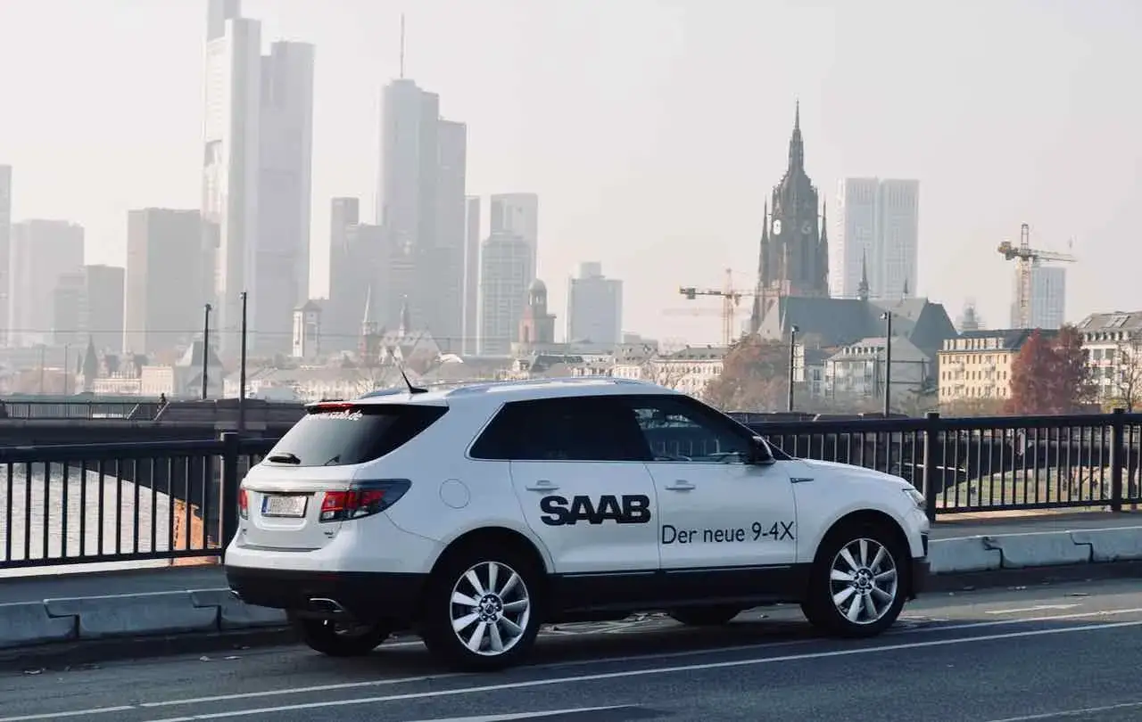 Vit Saab 9-4X parkerad p&aring; en bro med en stadssilhuett i bakgrunden.