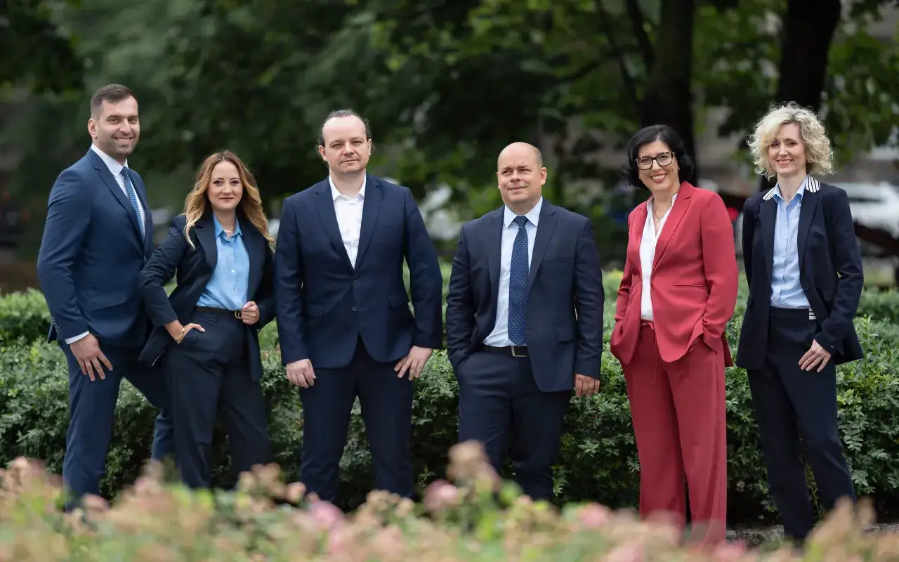 Grupa pięciu profesjonalistów w garniturach stoi na tle zieleni.