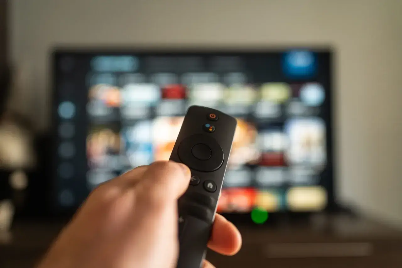Dłoń trzymająca pilot do telewizora, na ekranie widoczne kolorowe ikony aplikacji, gotowe do wyboru ulubionego serialu na web tv.