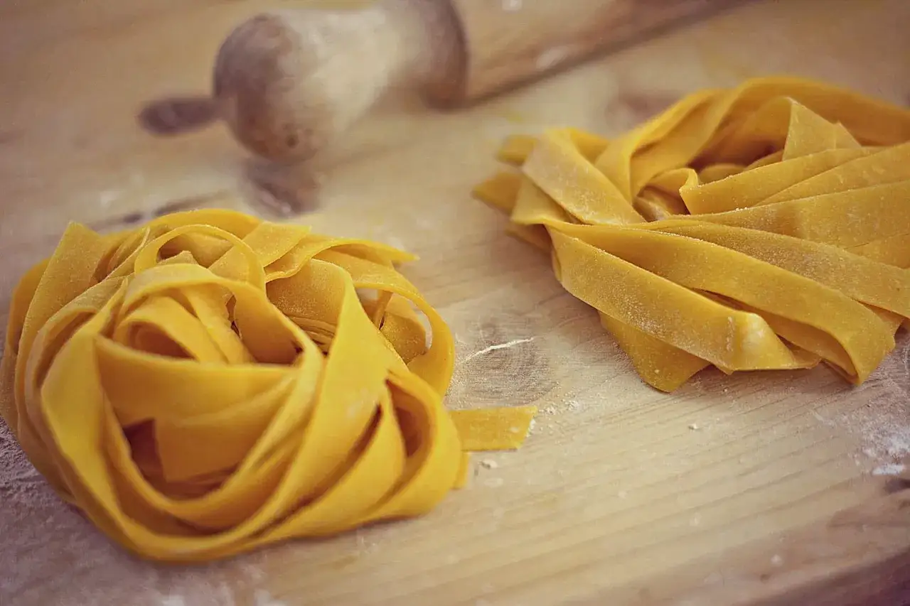 Fettuccine fresche su tagliere di legno, pronte per essere cucinate. Un assaggio di cosa mangiare a Forlì, con un mattarello sullo sfondo.