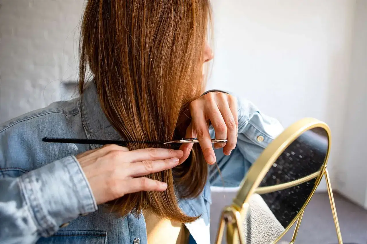 Frau schneidet sich die Haare zu Hause. Sie benutzt eine Schere und einen Kamm, um ihre braunen Haare zu stylen.
