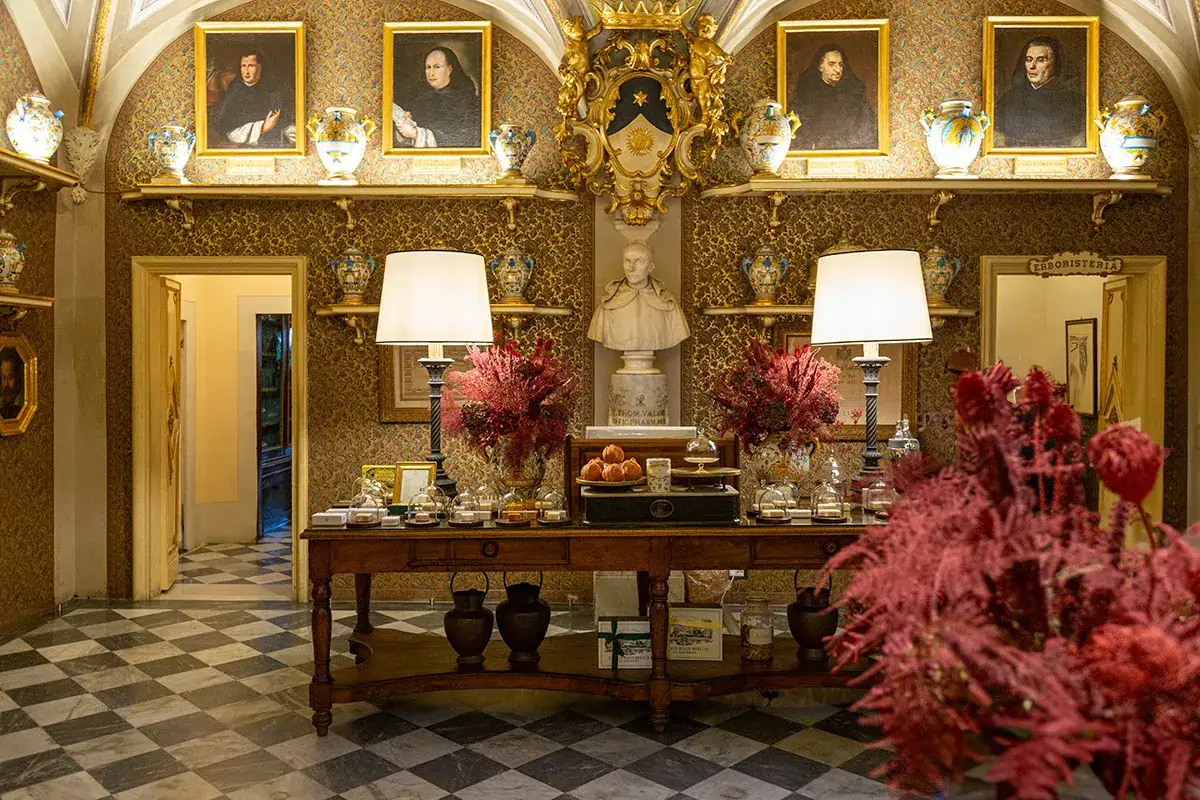 Interno elegante con ritratti antichi, vasi decorati e un tavolo con prodotti. L'atmosfera evoca la più antica farmacia di Firenze.