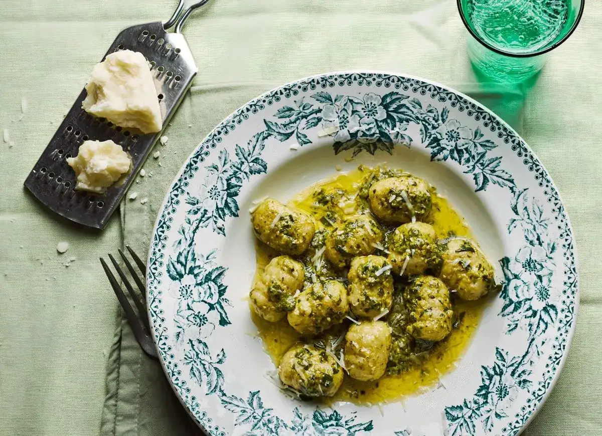Pyszne gnocchi z pesto, polane oliwą i posypane parmezanem, na talerzu z zielonym wzorem. Obok tarka z kawałkiem sera.