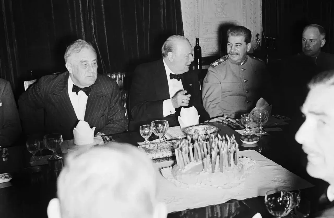 Roosevelt, Churchill y Stalin en la conferencia de Teherán, sentados a una mesa con un pastel de cumpleaños y velas encendidas.