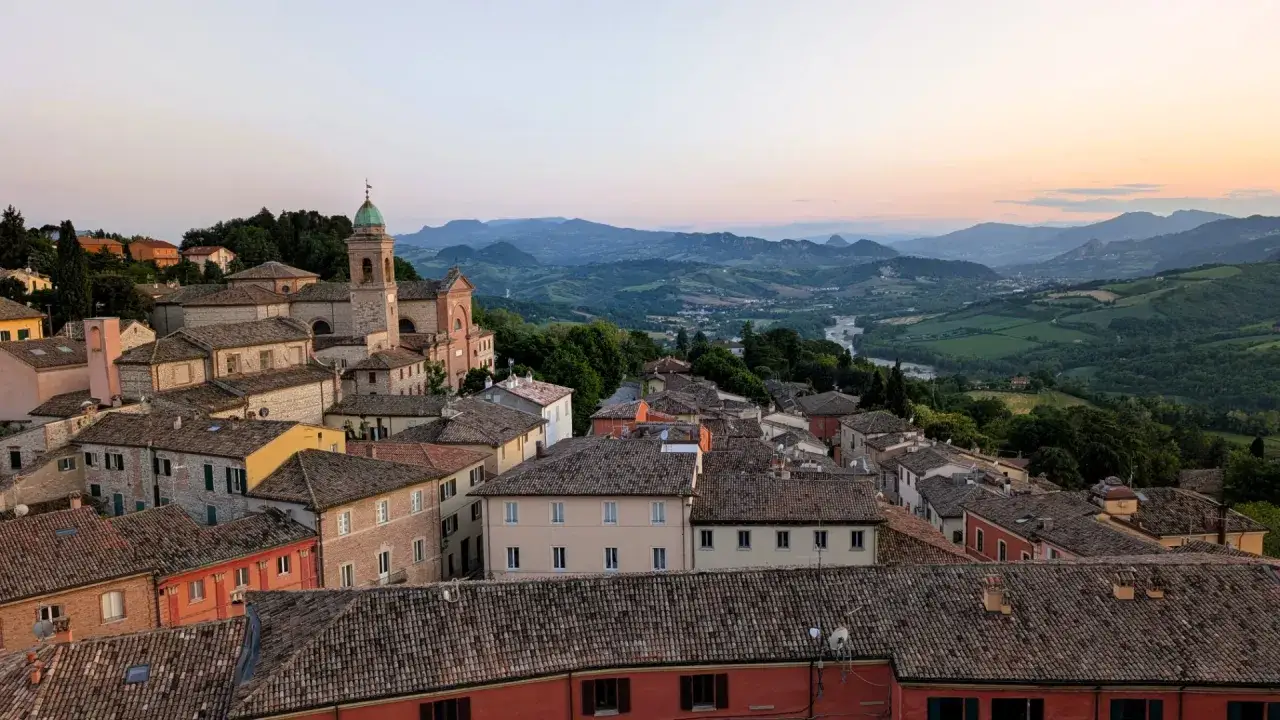 Panoramica di un borgo con tetti in cotto e una chiesa, incastonato tra le dolci colline riminesi al tramonto.