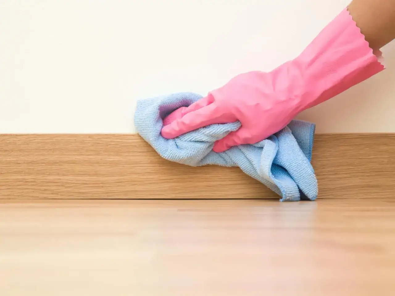 Mano con guante rosa limpiando rodapiés de madera con paño azul.