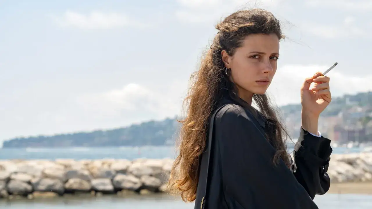 Ragazza con capelli lunghi e mossi, fuma una sigaretta, con sfondo di mare e costa. Il cielo &egrave; celeste dalla porta.
