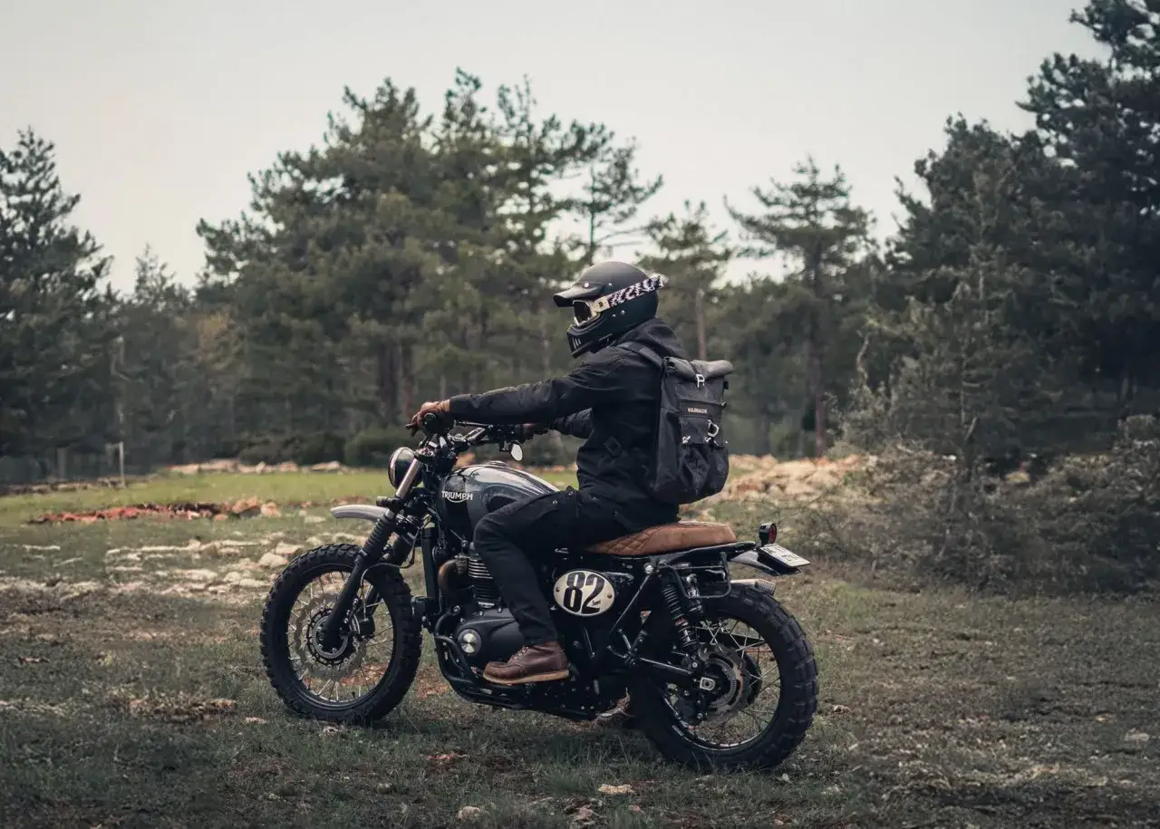 Motocyklista w kasku i plecaku na motorze scrambler, gotowy na przygodę w lesie.