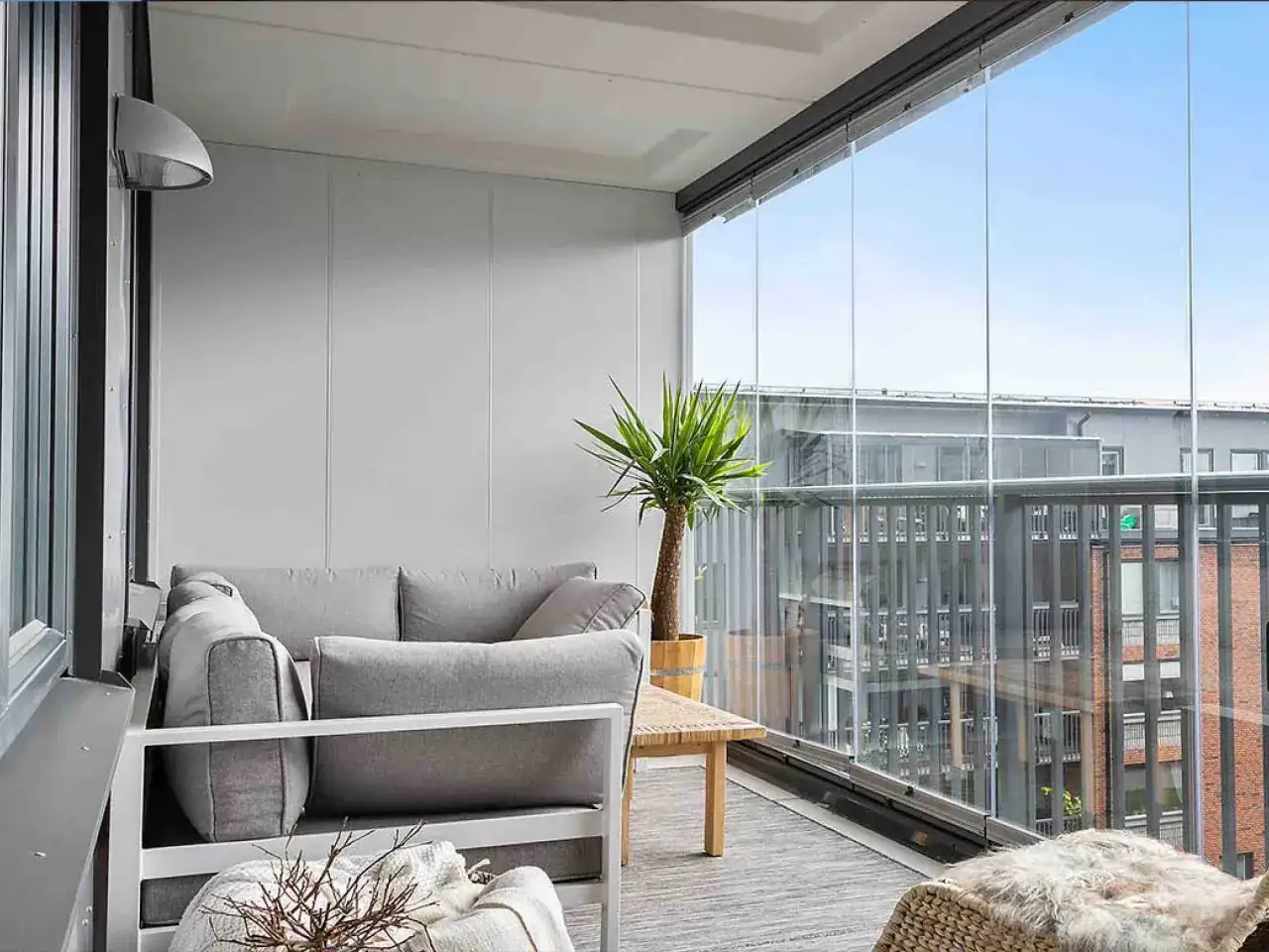Accogliente veranda balcone con divano grigio, tavolino in legno e pianta. Vetrate panoramiche offrono vista su edifici circostanti.