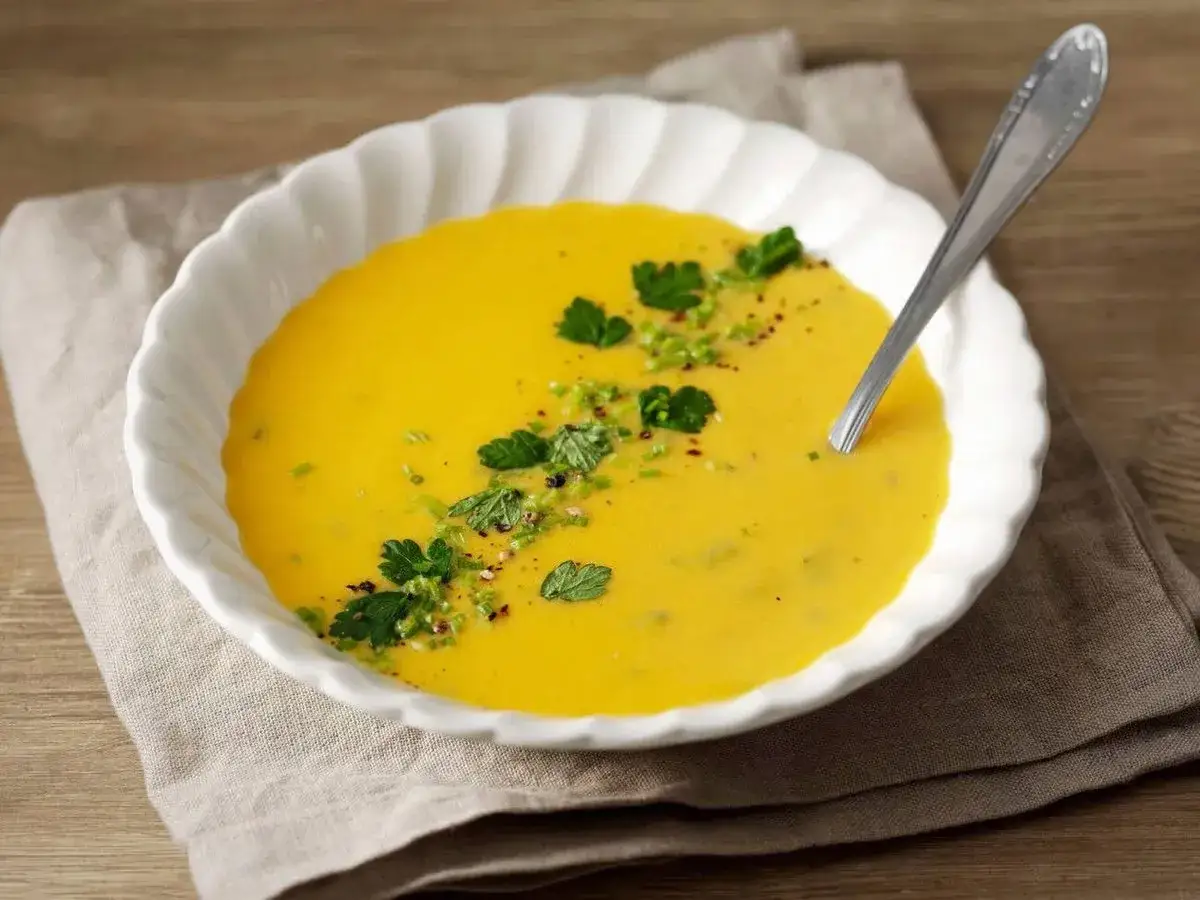 En kr&auml;mig morotssoppa med en klick creme fraiche, garnerad med f&auml;rska &ouml;rter och peppar. Serveras i en vit sk&aring;l med en sked.