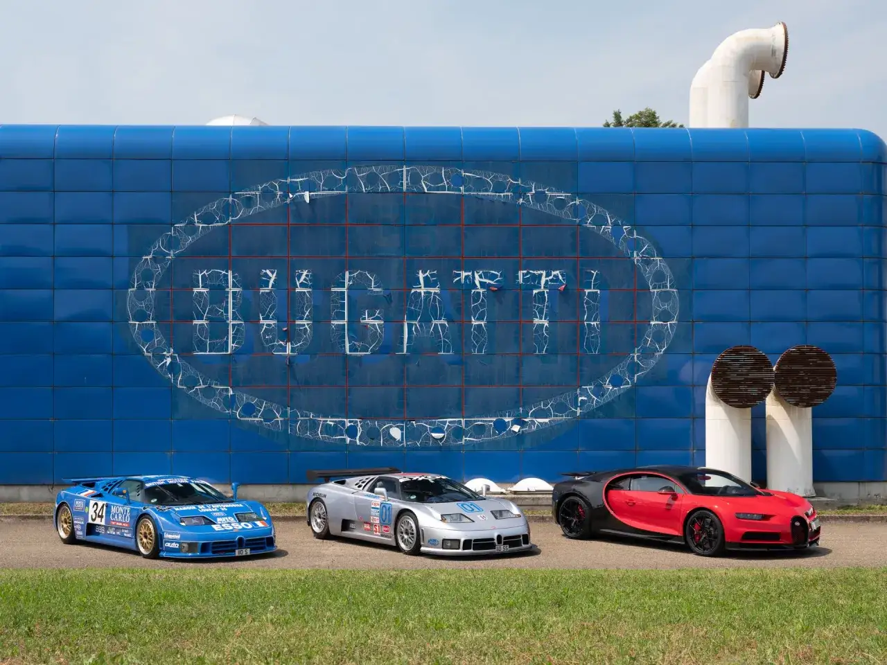 Trzy Bugatti, w tym niebieski bolid z numerem 34, srebrny z 01 i czerwono-czarny Chiron, na tle niebieskiej ściany z logo Bugatti. Z jakiego kraju jest Bugatti? Z Francji!