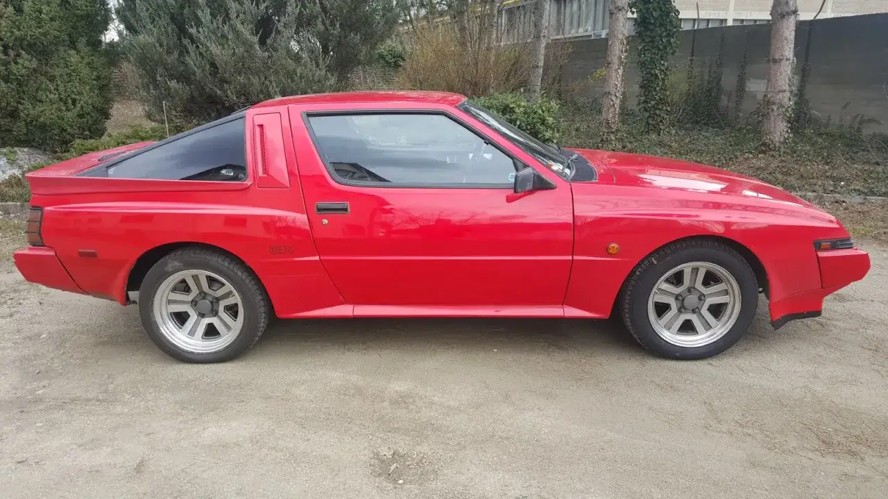 Czerwony Mitsubishi Starion EX z lat 80. stoi na żwirowej drodze, otoczony zielenią i drzewami.