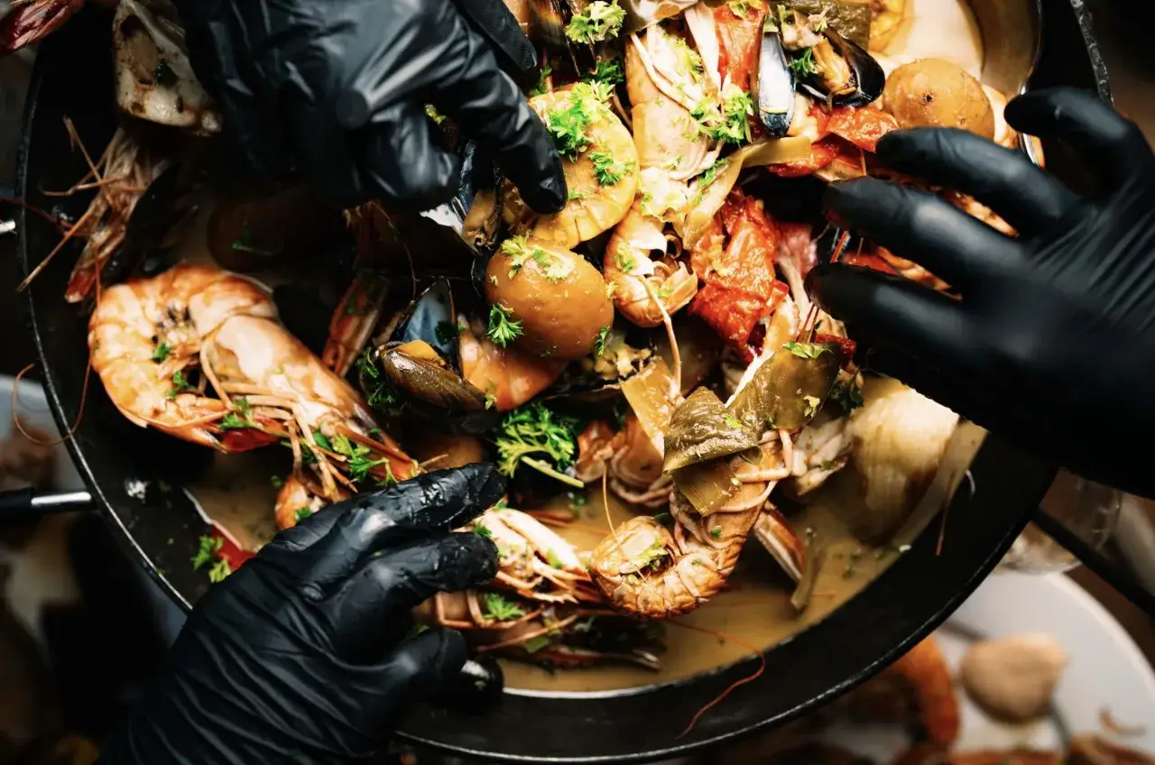 Händer i svarta handskar plockar ur en stor panna med en härlig seafood gastro-mix av räkor, musslor och kräftor, garnerad med persilja.