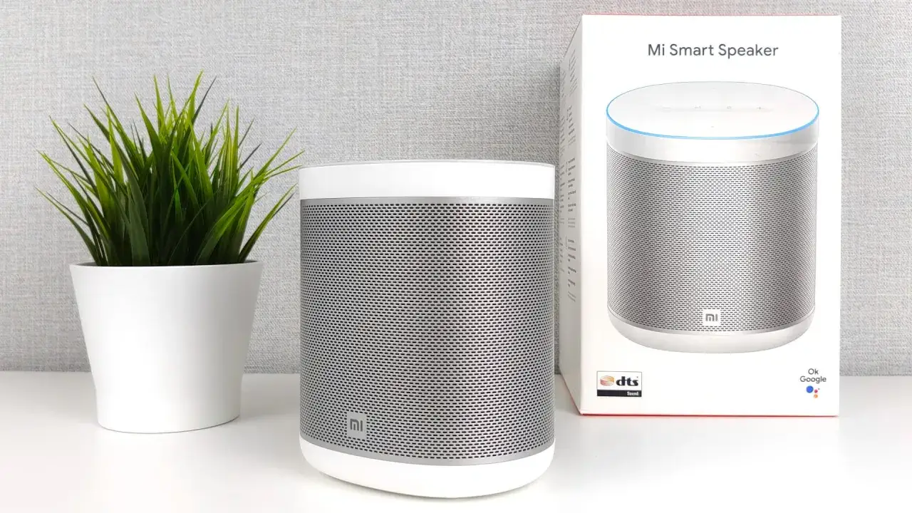 Ein wei&szlig;er Mi Smart Speaker steht neben seiner Verpackung. Eine gr&uuml;ne Pflanze im Topf steht links daneben.