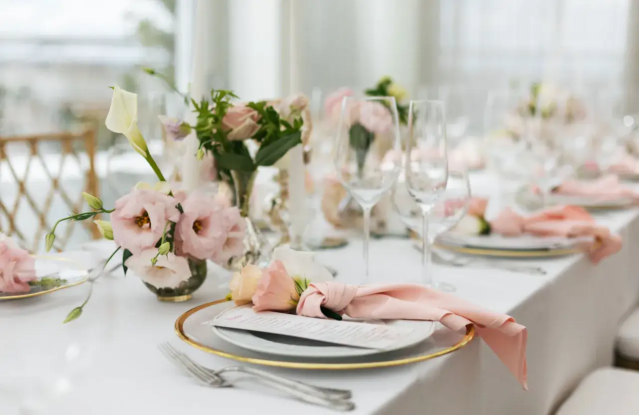 En vacker dukning med rosa blommor, guldkantade tallrikar och ljusa servetter. Perfekt för ett elegant firande.