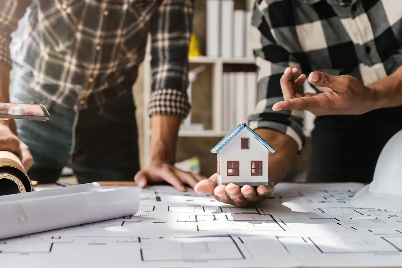 Dwóch mężczyzn omawia projektowanie domu online, trzymając w ręku model domu na tle planów architektonicznych.