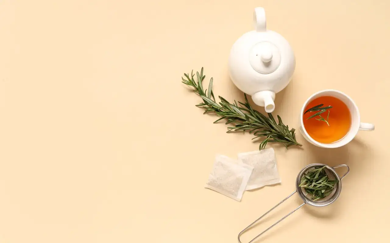 Une théière blanche, une tasse de tisane romarin, des sachets et une passoire remplie de romarin frais sur fond beige.