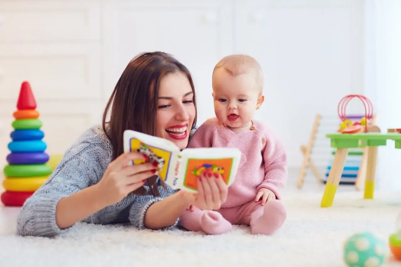 Mama czyta książeczkę 20 miesięcznemu dziecku. Radosna atmosfera, kolorowe zabawki w tle.