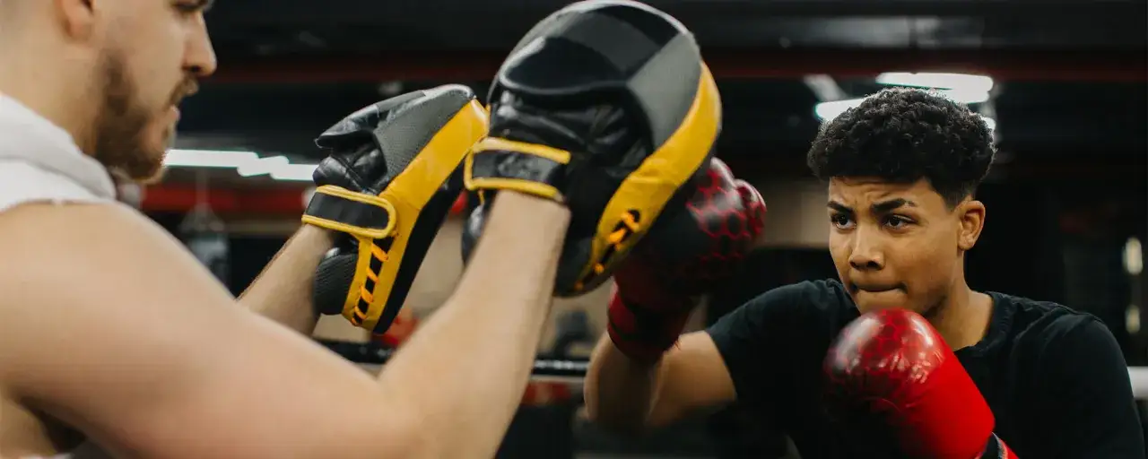 A boxer, possibly a southpaw, trains with a coach holding pads. The boxer wears red gloves, focused and ready.