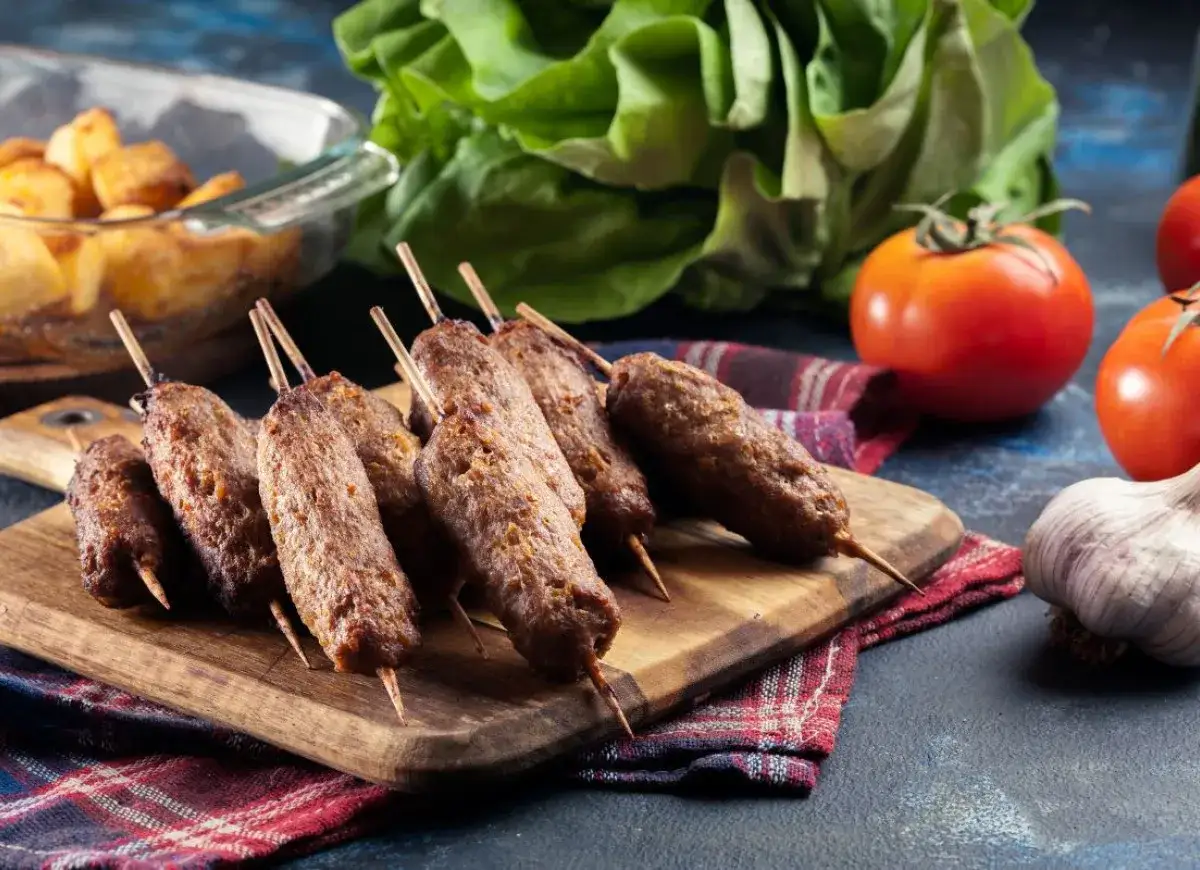 Pyszne, grillowane ćevapi na drewnianej desce, obok pomidorów, czosnku i sałaty.