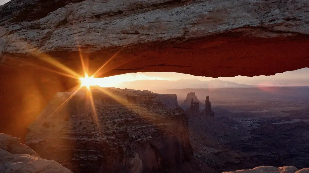 Lever du soleil à travers une arche rocheuse, avec un effet de lens flare créé par Photoshop, illuminant le paysage désertique.