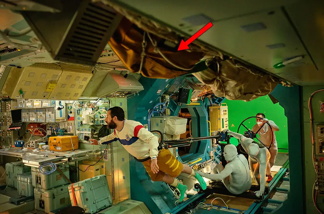 Scena z planu filmowego, gdzie aktor w stroju kosmicznym przypomina Adama Sandlera. W tle zielone tło i ekipa techniczna.