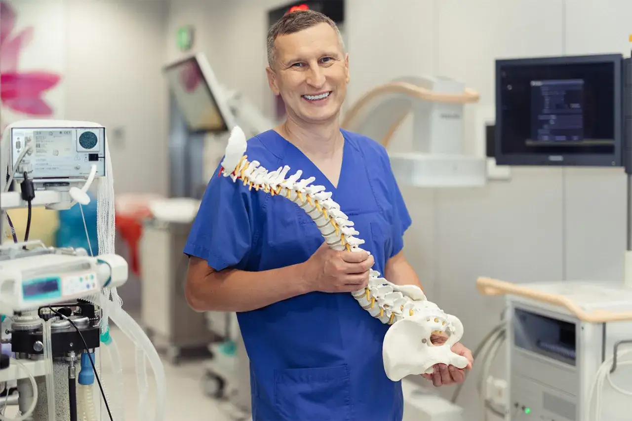 Uśmiechnięty lekarz w niebieskim uniformie trzyma model kręgosłupa. To jeden z najlepszych neurochirurgów kręgosłupa w Polsce, gotowy do pomocy.
