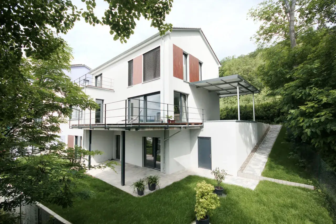 Modernes Haus mit Balkonen und &uuml;berdachter Terrasse. Ein schmaler Weg f&uuml;hrt den Hang hinauf, umgeben von Gr&uuml;n. Der spritzschutz am Haus ist dezent integriert.