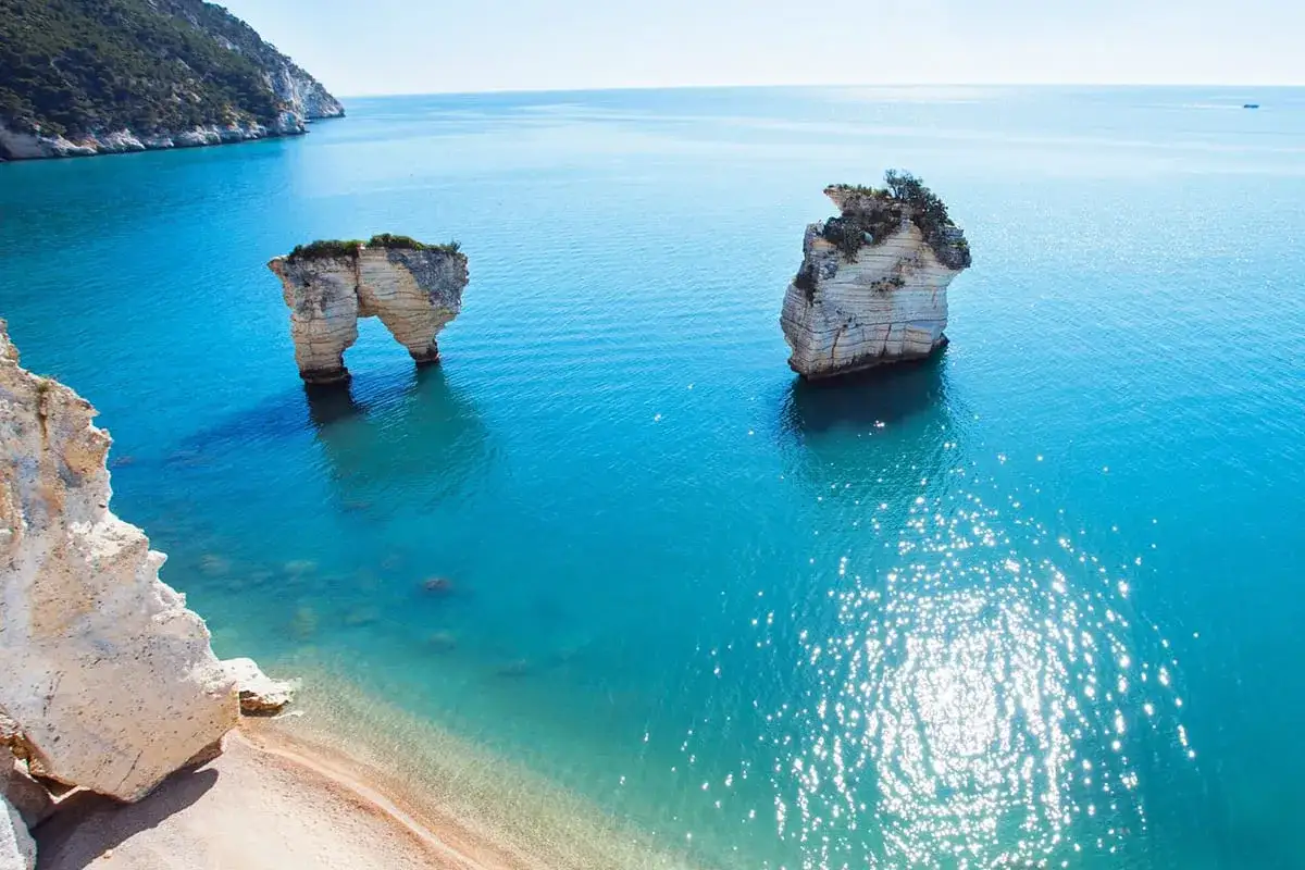 La spiaggia di Piana di Mattinata, con le sue iconiche rocce a forma di arco e isolotti, bagnata da un mare cristallino e soleggiato.